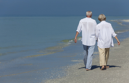 Rear view of happy senior man and woman couple walking and holding hands on a deserted tropical beach with bright clear blue sky